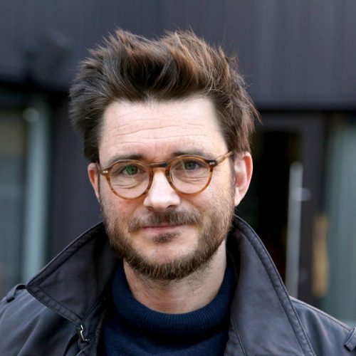 A man in a dark blue coat looks at the camera. He has round glasses, a beard and brown hair.